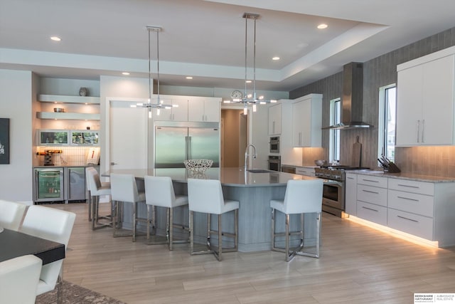 kitchen featuring hanging light fixtures, wall chimney range hood, premium appliances, a kitchen island with sink, and white cabinets