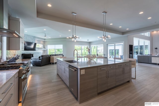 kitchen with sink, wall chimney exhaust hood, a large island with sink, appliances with stainless steel finishes, and light wood-type flooring