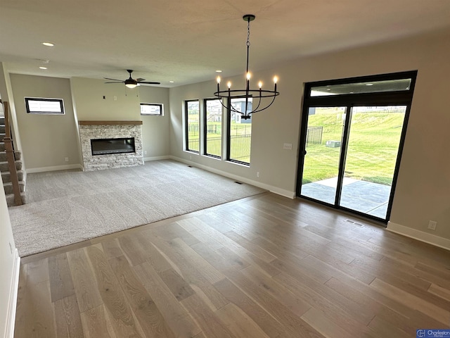 unfurnished living room featuring ceiling fan with notable chandelier, hardwood / wood-style flooring, a stone fireplace, and a wealth of natural light