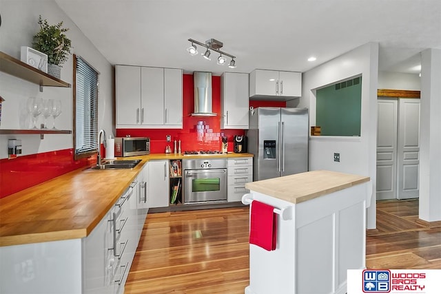 kitchen featuring butcher block counters, sink, wall chimney range hood, white cabinets, and appliances with stainless steel finishes