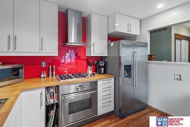 kitchen featuring wooden counters, white cabinets, wall chimney range hood, appliances with stainless steel finishes, and wood-type flooring