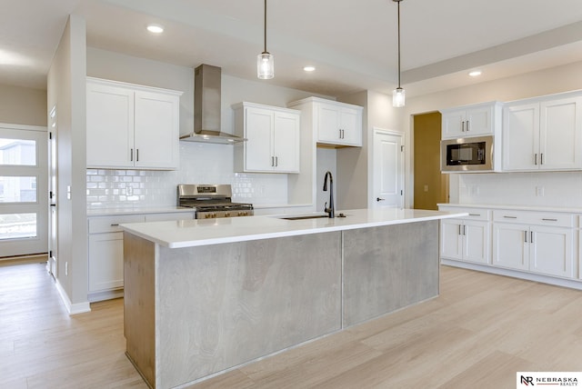 kitchen featuring light wood-style flooring, a sink, wall chimney range hood, appliances with stainless steel finishes, and a center island with sink