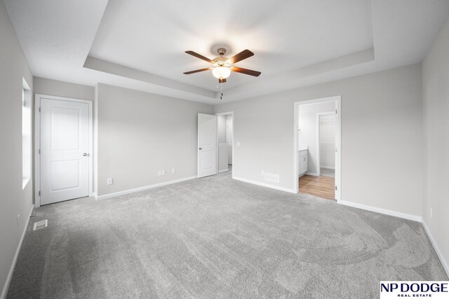 unfurnished bedroom with a raised ceiling, connected bathroom, ceiling fan, and light colored carpet