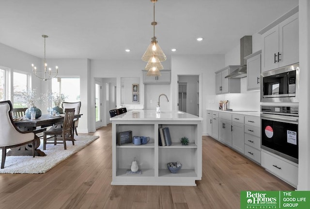 kitchen featuring pendant lighting, a center island with sink, light hardwood / wood-style flooring, wall chimney exhaust hood, and appliances with stainless steel finishes