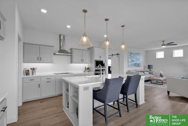 kitchen with wall chimney range hood, a barn door, light hardwood / wood-style floors, a kitchen island with sink, and a breakfast bar