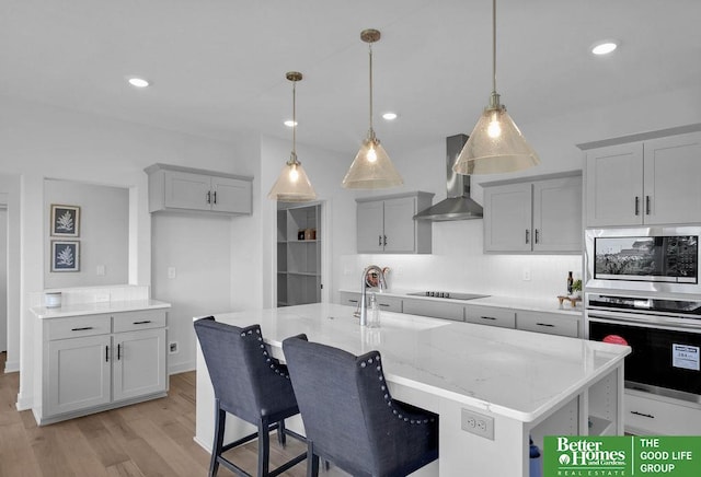 kitchen with gray cabinetry, wall chimney range hood, light hardwood / wood-style flooring, an island with sink, and stainless steel appliances