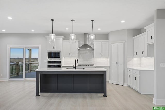 kitchen with stainless steel appliances, light hardwood / wood-style floors, a center island with sink, and wall chimney range hood