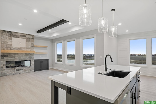 kitchen with pendant lighting, a stone fireplace, sink, an island with sink, and light hardwood / wood-style floors