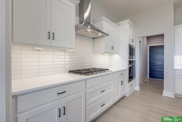 kitchen featuring stainless steel appliances, wall chimney range hood, backsplash, white cabinets, and light wood-type flooring