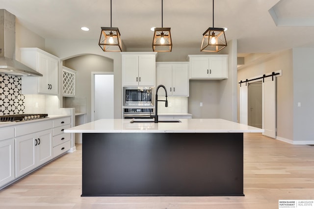 kitchen featuring stainless steel appliances, wall chimney range hood, a barn door, hanging light fixtures, and an island with sink