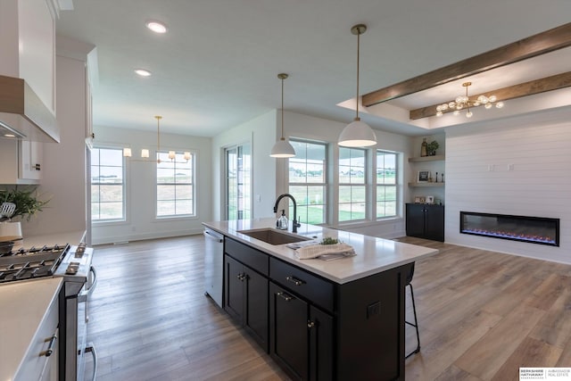 kitchen with wall chimney exhaust hood, stainless steel appliances, a kitchen island with sink, an inviting chandelier, and white cabinets