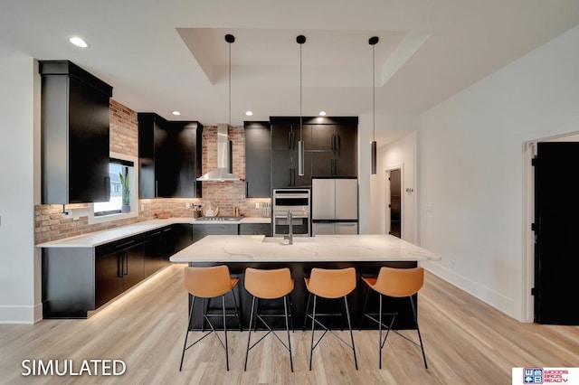 kitchen with a center island with sink, wall chimney exhaust hood, stainless steel appliances, pendant lighting, and a raised ceiling