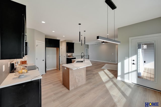 kitchen with a kitchen island with sink, hanging light fixtures, light hardwood / wood-style flooring, ceiling fan, and light stone countertops