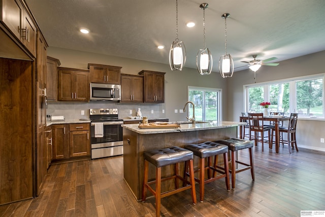 kitchen with a kitchen island with sink, light stone countertops, dark hardwood / wood-style floors, and appliances with stainless steel finishes