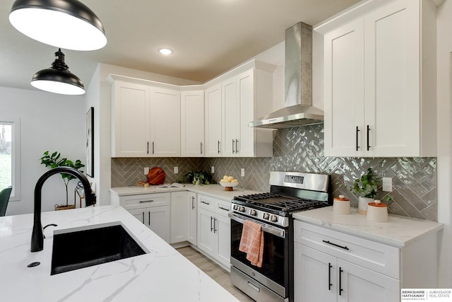 kitchen with light stone counters, stainless steel range with gas cooktop, sink, wall chimney range hood, and white cabinetry