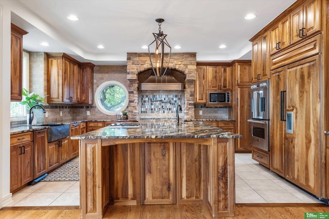 kitchen with light wood-type flooring, built in appliances, a center island with sink, and dark stone counters