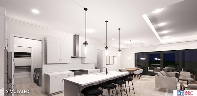 kitchen with light wood-type flooring, a kitchen island with sink, wall chimney range hood, decorative light fixtures, and white cabinetry