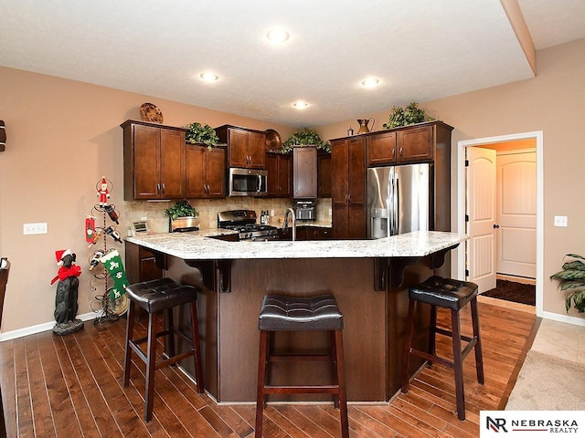 kitchen with dark wood-type flooring, tasteful backsplash, appliances with stainless steel finishes, a kitchen breakfast bar, and dark brown cabinetry