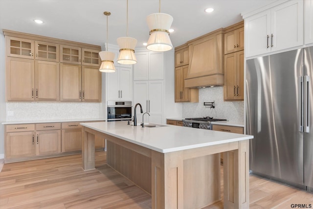 kitchen featuring a kitchen island with sink, light hardwood / wood-style flooring, appliances with stainless steel finishes, decorative light fixtures, and custom range hood