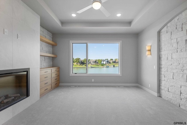 unfurnished living room with ceiling fan, light colored carpet, a water view, and a tray ceiling