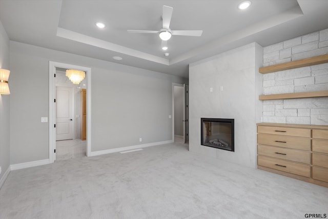 unfurnished living room featuring a raised ceiling, ceiling fan, a fireplace, and light colored carpet