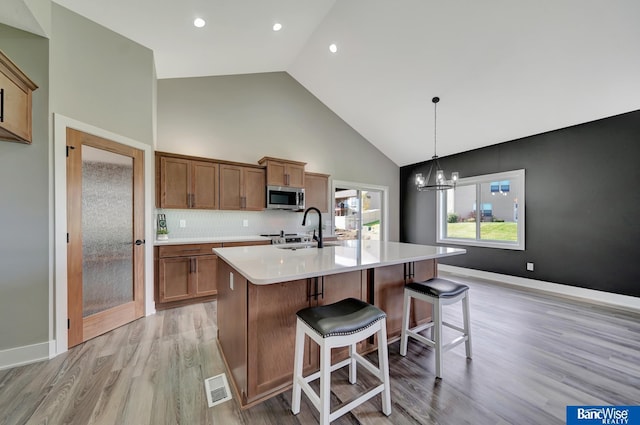 kitchen featuring a kitchen island with sink, high vaulted ceiling, hanging light fixtures, light wood-type flooring, and tasteful backsplash