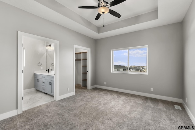 unfurnished bedroom with light carpet, a walk in closet, a raised ceiling, ceiling fan, and connected bathroom