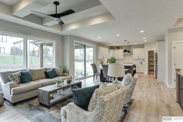 living room with plenty of natural light, ceiling fan, a raised ceiling, and light wood-type flooring