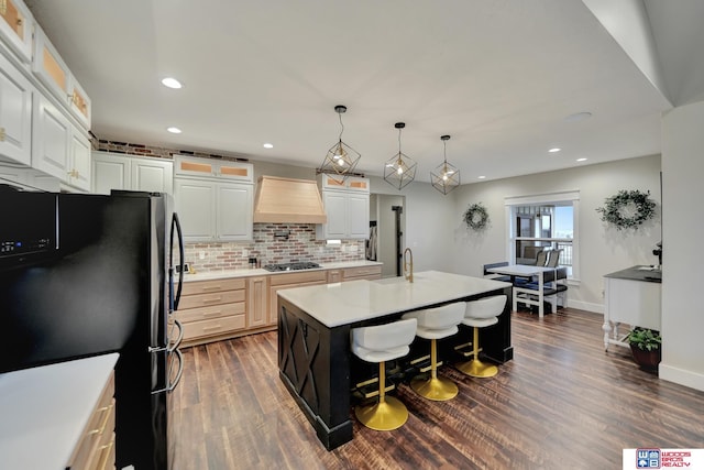 kitchen featuring custom exhaust hood, a center island with sink, black fridge, decorative light fixtures, and stainless steel gas cooktop