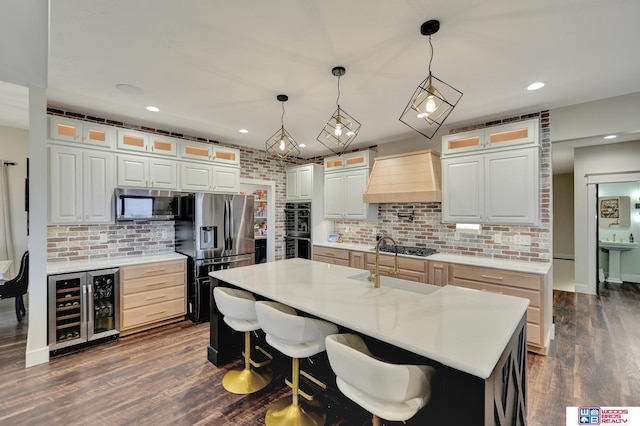 kitchen with custom exhaust hood, a kitchen island with sink, hanging light fixtures, wine cooler, and stainless steel appliances