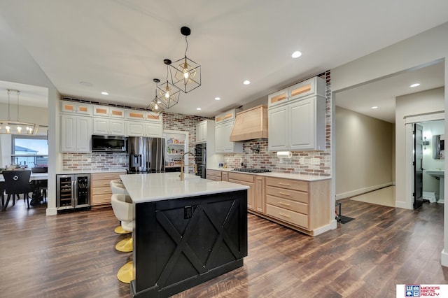 kitchen with hanging light fixtures, wine cooler, a kitchen island with sink, custom range hood, and appliances with stainless steel finishes