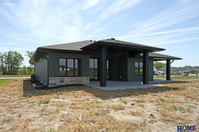 exterior space with stone siding, roof with shingles, central AC unit, and a patio