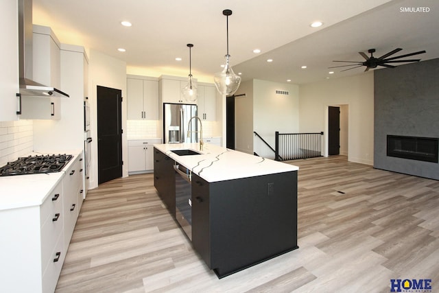 kitchen featuring white cabinets, an island with sink, wall chimney exhaust hood, appliances with stainless steel finishes, and decorative light fixtures