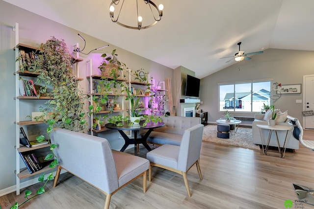 dining room featuring baseboards, light wood-type flooring, vaulted ceiling, ceiling fan with notable chandelier, and a glass covered fireplace