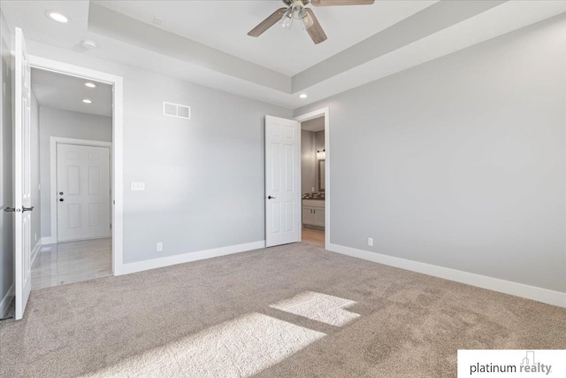 unfurnished bedroom with a tray ceiling, connected bathroom, ceiling fan, and light colored carpet