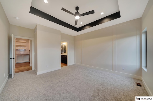 unfurnished bedroom featuring a tray ceiling, ensuite bath, ceiling fan, and light carpet