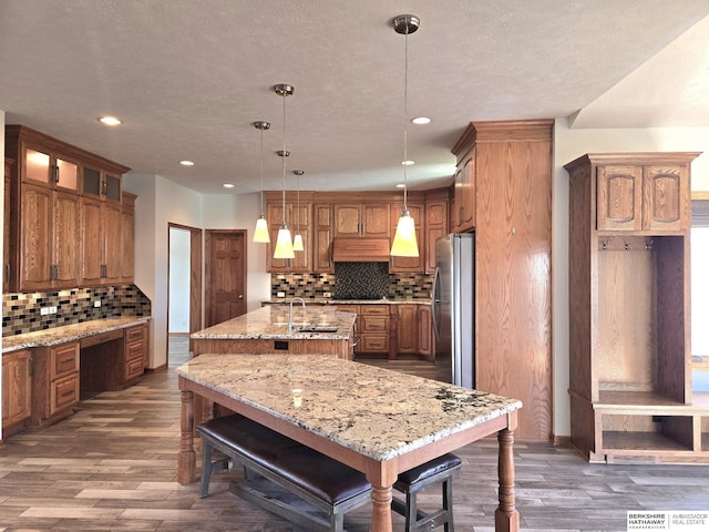 kitchen featuring stainless steel fridge, tasteful backsplash, custom exhaust hood, a kitchen island with sink, and hanging light fixtures