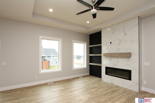 unfurnished living room with ceiling fan, a raised ceiling, built in features, a high end fireplace, and light hardwood / wood-style flooring