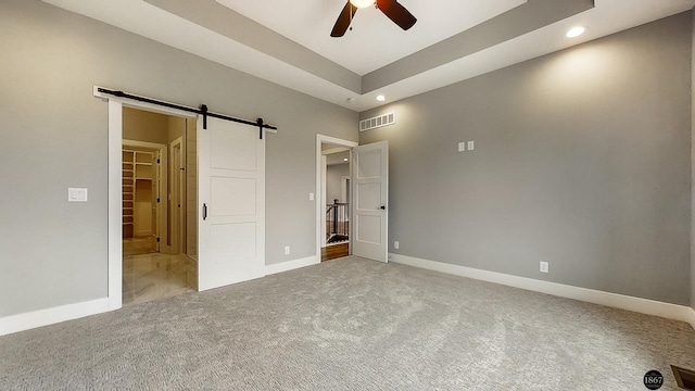 unfurnished bedroom featuring a walk in closet, a raised ceiling, ceiling fan, a barn door, and carpet floors