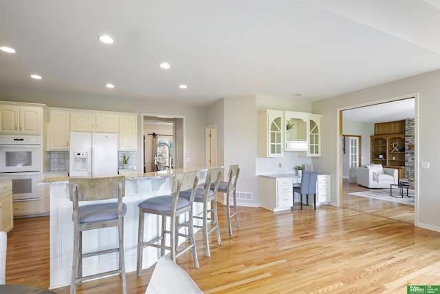 kitchen featuring decorative backsplash, a kitchen breakfast bar, white appliances, a kitchen island with sink, and light hardwood / wood-style floors