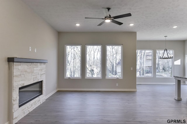 unfurnished living room featuring a stone fireplace, a wealth of natural light, dark wood-type flooring, and ceiling fan with notable chandelier