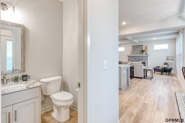 bathroom featuring beam ceiling, a stone fireplace, hardwood / wood-style floors, toilet, and vanity