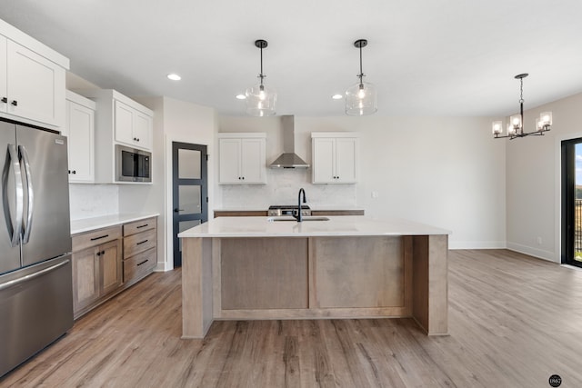 kitchen featuring white cabinets, a center island with sink, wall chimney range hood, sink, and stainless steel refrigerator