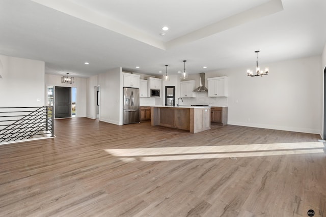 kitchen with a spacious island, wall chimney range hood, hanging light fixtures, stainless steel fridge, and white cabinetry