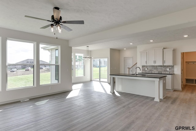 kitchen with decorative backsplash, ceiling fan with notable chandelier, a center island with sink, light hardwood / wood-style flooring, and white cabinetry