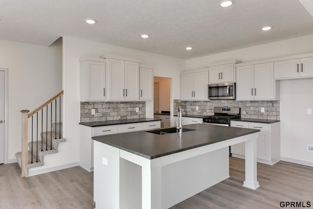 kitchen with a kitchen island with sink, sink, light wood-type flooring, appliances with stainless steel finishes, and white cabinetry