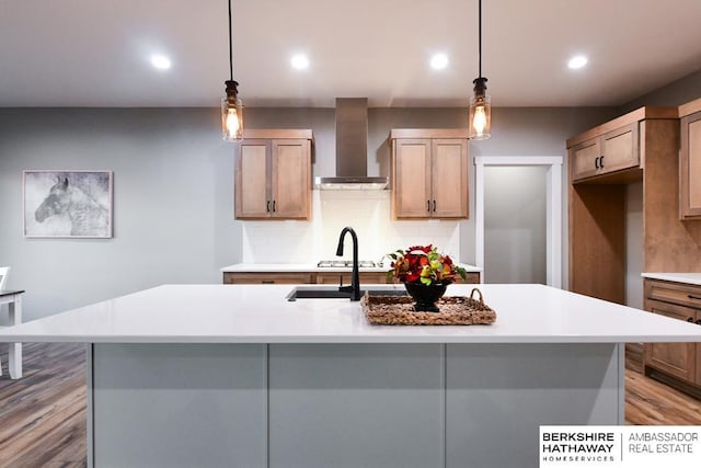 kitchen with wall chimney range hood, an island with sink, hanging light fixtures, and tasteful backsplash