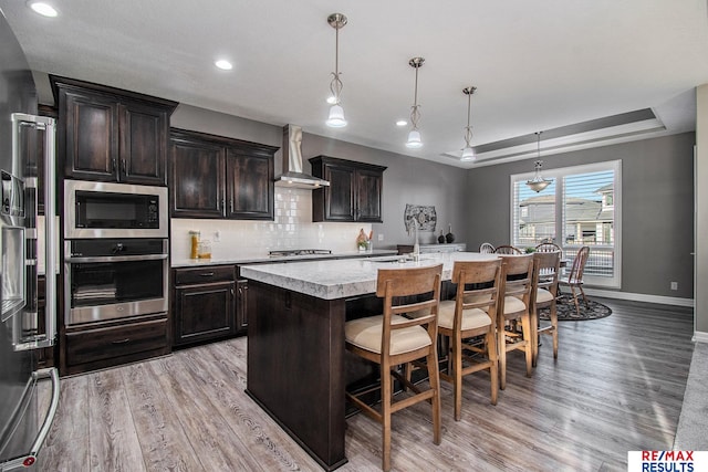 kitchen featuring a kitchen island with sink, pendant lighting, stainless steel appliances, and wall chimney range hood