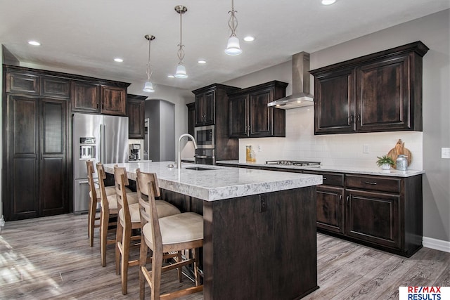 kitchen featuring appliances with stainless steel finishes, wall chimney exhaust hood, a center island with sink, light hardwood / wood-style floors, and hanging light fixtures