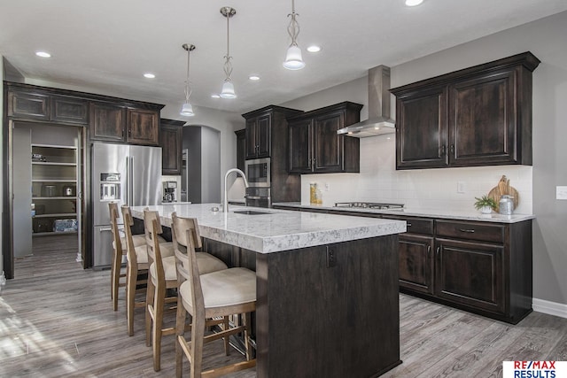 kitchen with sink, wall chimney exhaust hood, decorative light fixtures, a kitchen island with sink, and appliances with stainless steel finishes